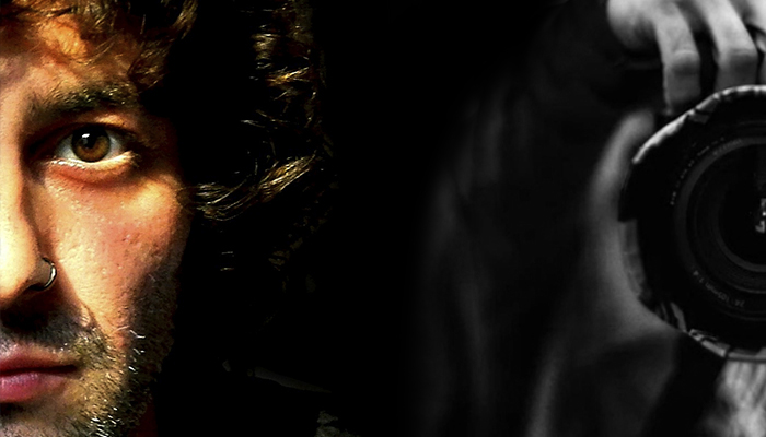 Fotografía en primer plano del objetivo de la cámara y mano del fotógrafo realizada frente a un espejo. El punto de enfoque permite ver ese reflejo mientras el fondo de la ciudad queda totalmente desenfocado. Se pueden apreciar reflejos de luces y poco más.